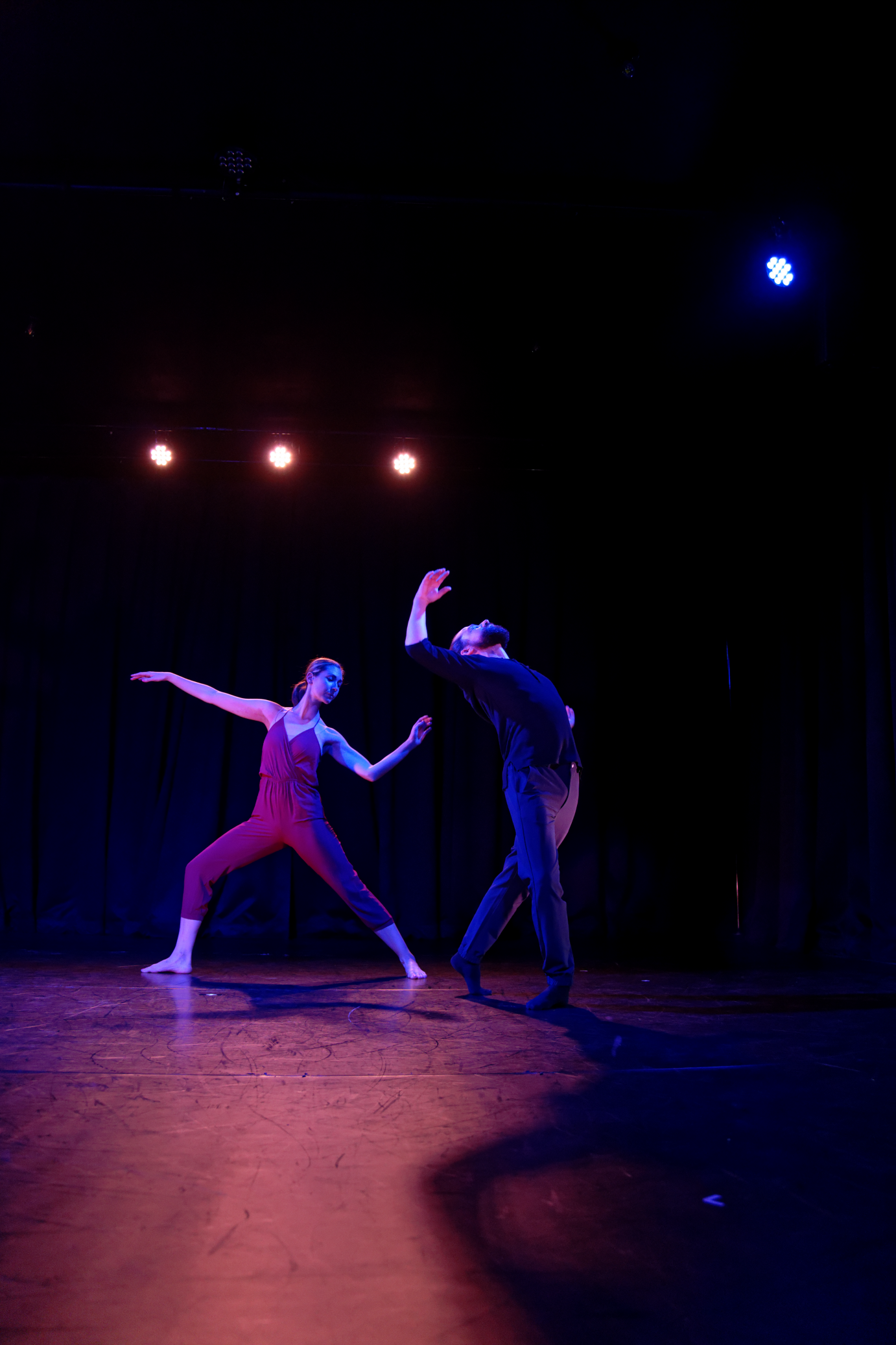 In a red unitard, Siobhan Murray lunges in fourth position. She lifts one arm to Aaron Selissen, who is backbending over her hand. One of his arms is curved and lifted.